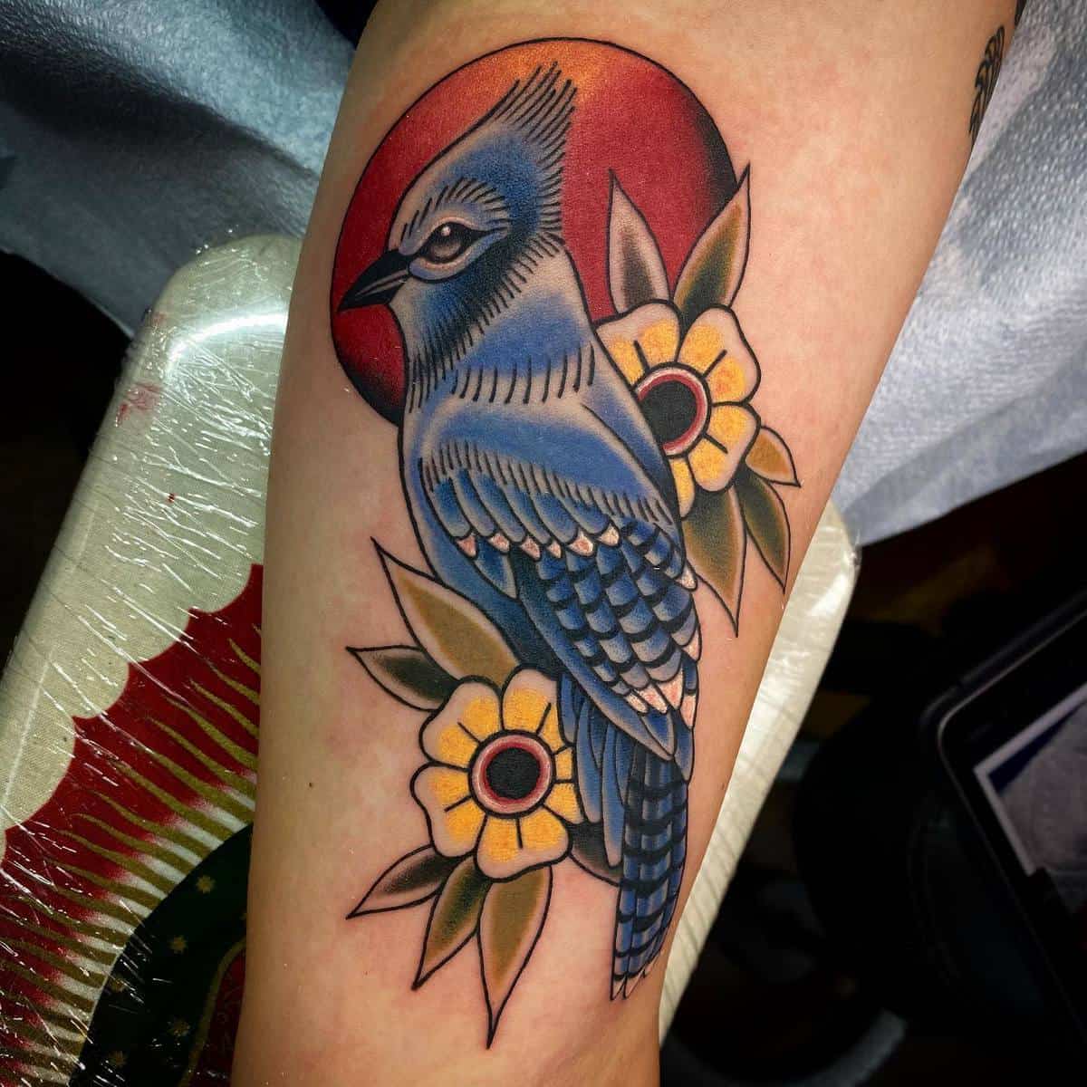 Tattoo of a blue bird with yellow flowers and a red sun background on an arm