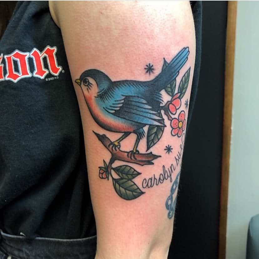 Tattoo of a colorful bird on a branch with leaves and flowers, with cursive text, on an upper arm