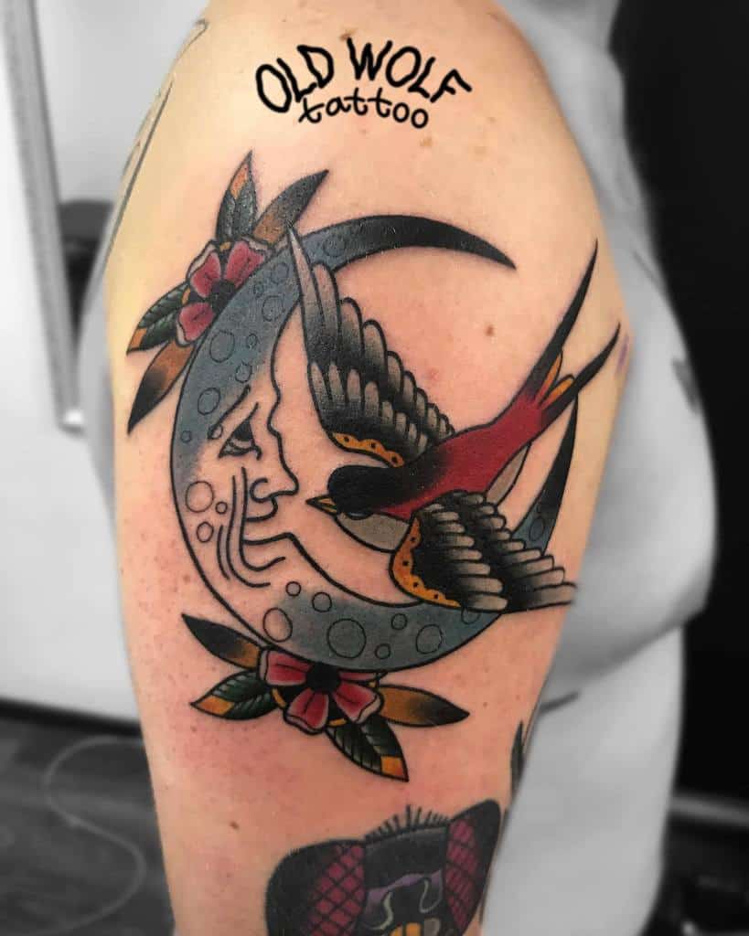 Arm tattoo of a flying bird over a crescent moon framed with flowers and a banner text