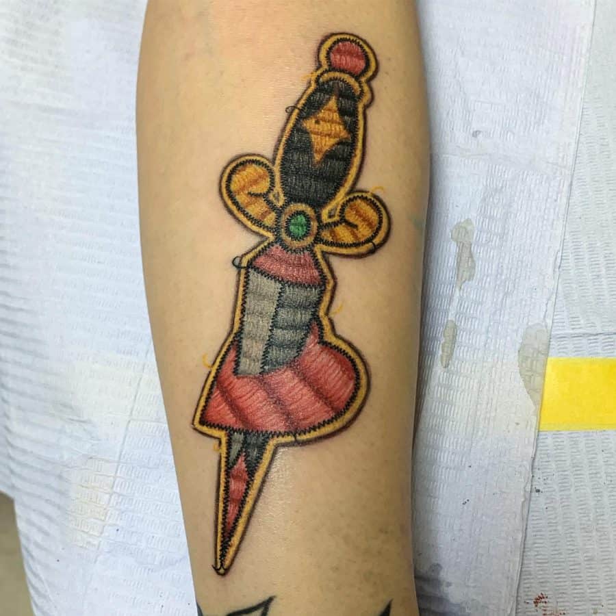 Patchwork-style tattoo of an antique dagger with vibrant embroidery details, giving it the appearance of a fabric appliqué.