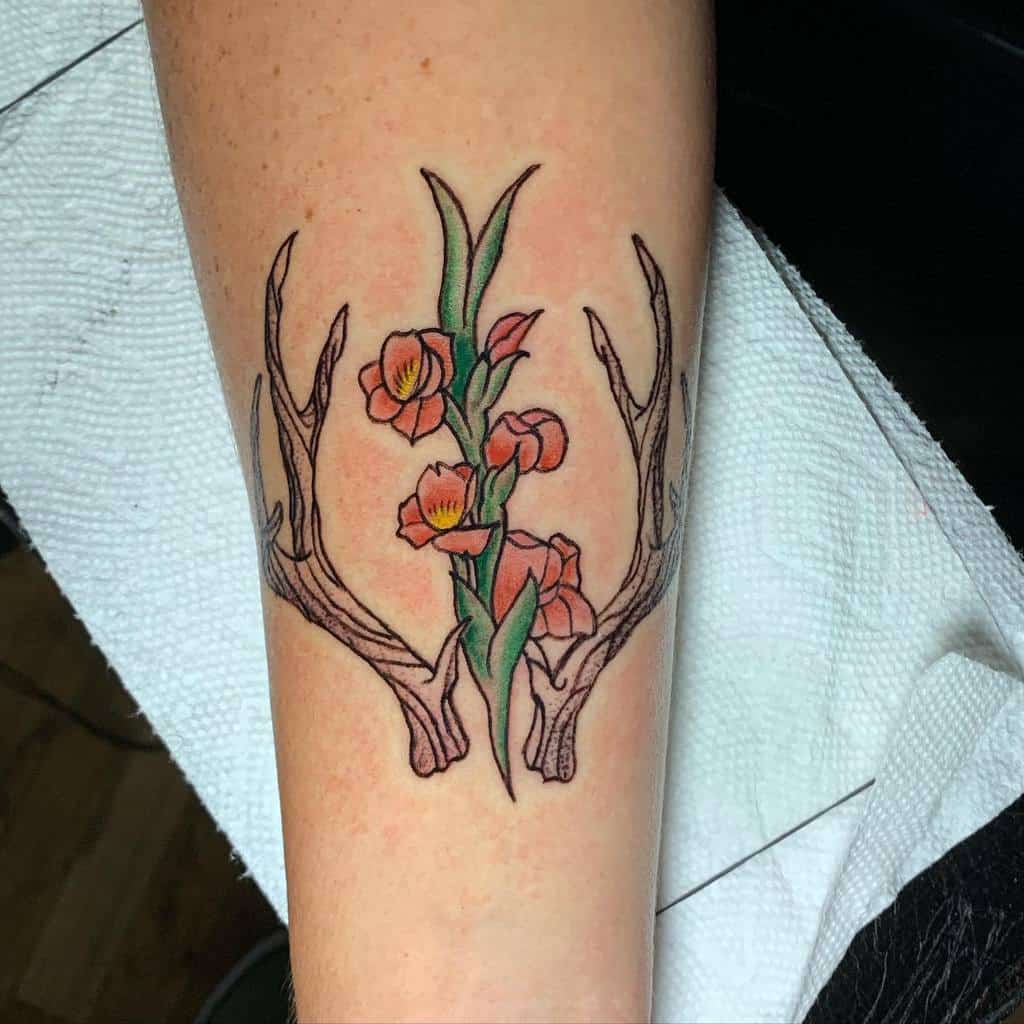 Tattoo of antlers intertwined with orange flowers, green leaves, and vibrant gladiolus on an arm