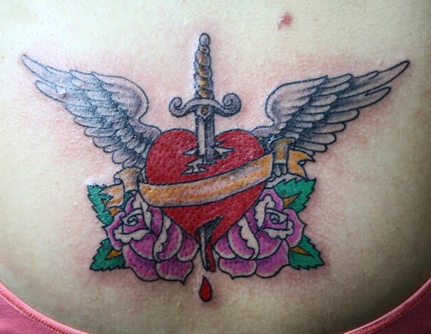 A tattoo of a heart with wings, pierced by a sword, adorned with a banner, and surrounded by roses below, all with blood dripping elegantly