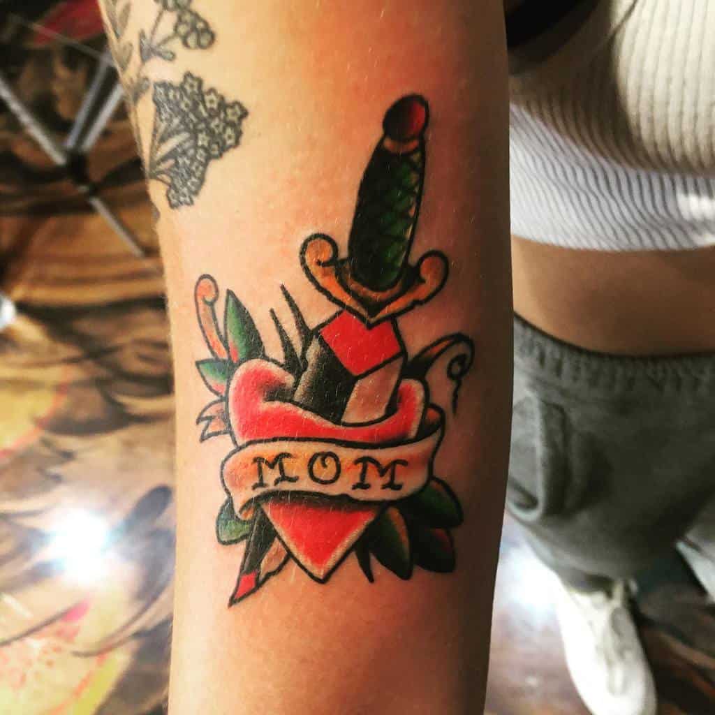 Tattoo of a dagger through a heart with a "MOM" banner on person's arm, honoring their love and dedication