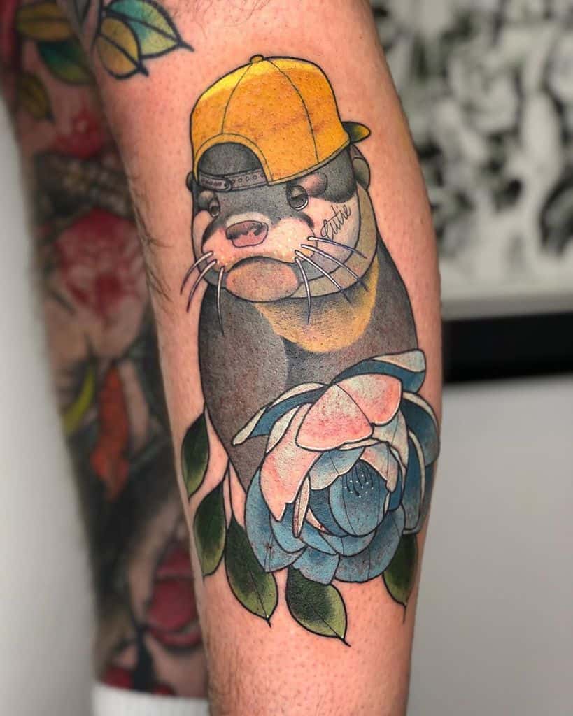 Tattoo of an otter wearing a yellow cap, sitting on a blue and green flower with leaves