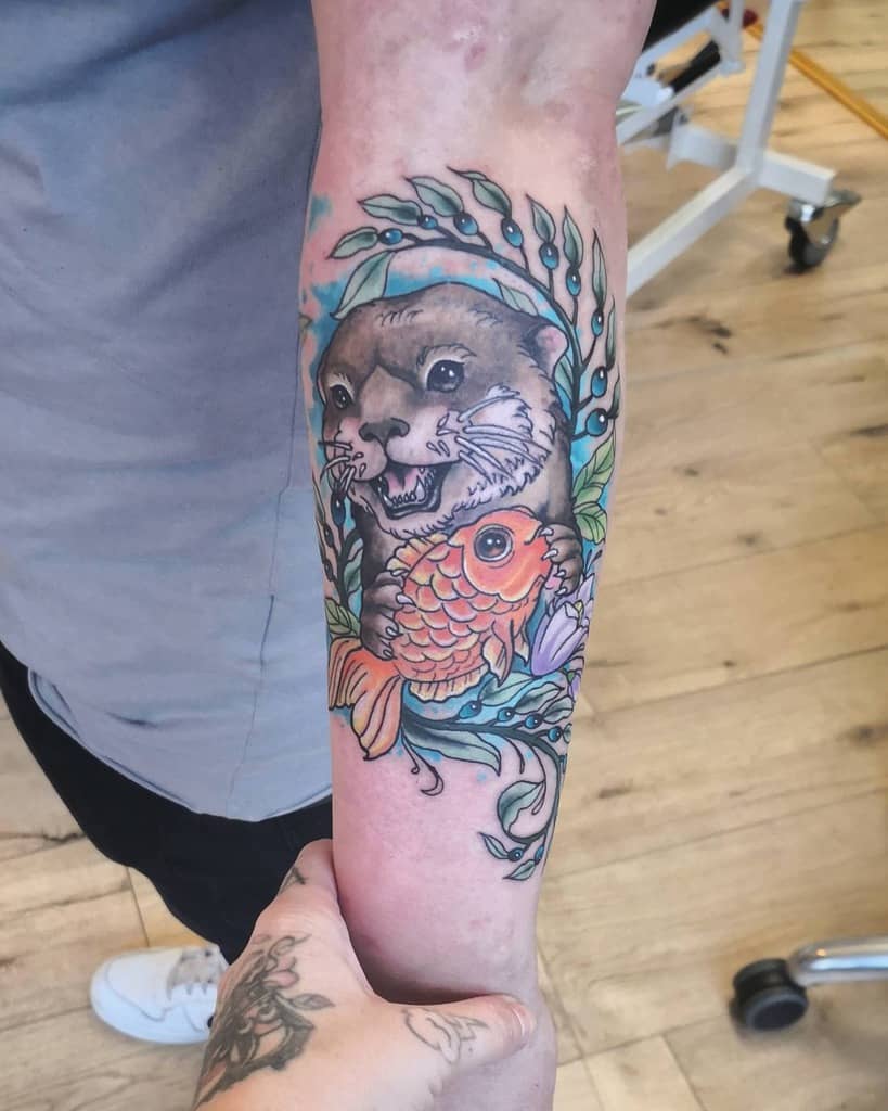 Forearm tattoo of an otter and colorful fish surrounded by leaves, with a hand holding the arm