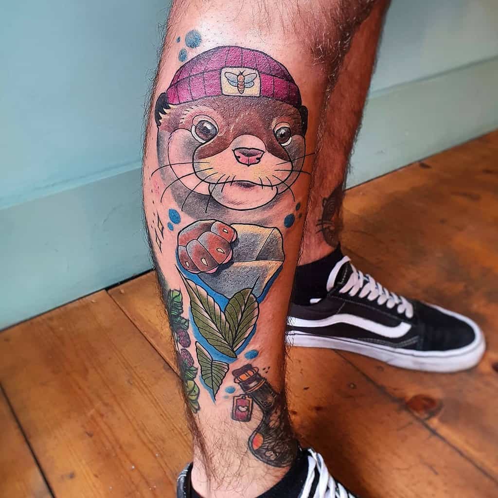 Colorful tattoo on a person's leg featuring an otter with a hat, a diamond, leaves, and various vibrant elements