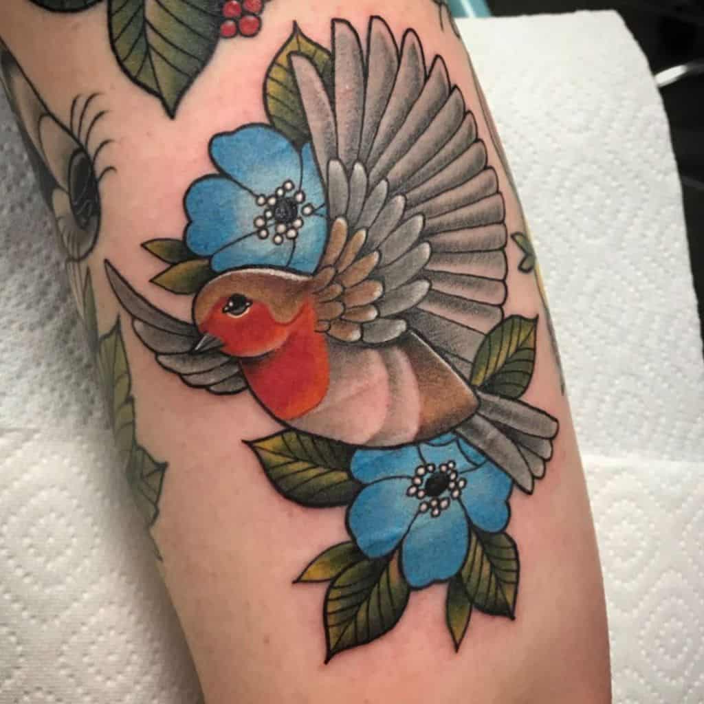 Tattoo of a robin with wings spread, surrounded by blue flowers and green leaves