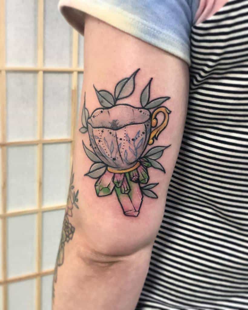 Colorful tattoo of a teacup with leafy details, featuring a golden handle and resting on vibrant pink and green crystals for a mystical touch, surrounded by leaves
