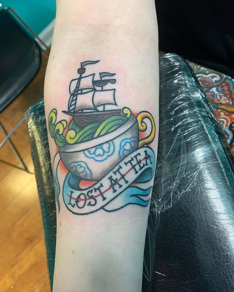Colorful tattoo of a ship in a blue floral teacup with swirling waves, accompanied by a ribbon reading "Lost at Tea" 