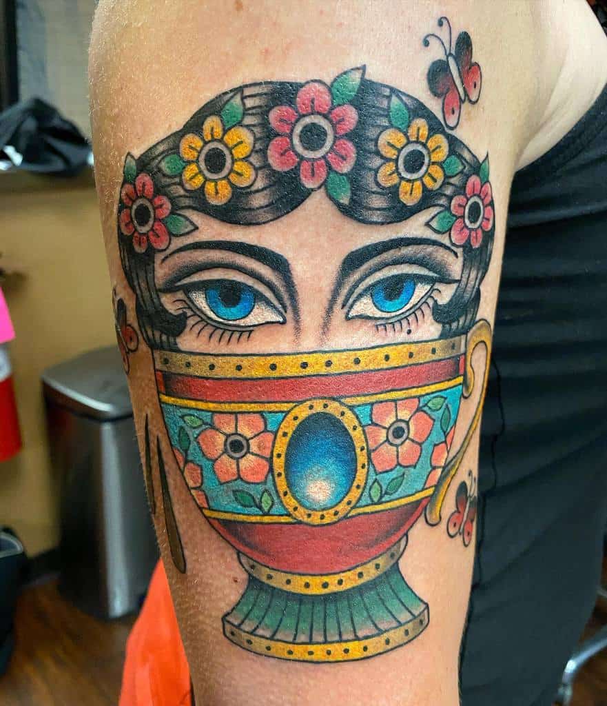 Colorful tattoo of a woman's face with blue eyes, her lower face hidden behind a colorful floral teacup, with a flower-adorned hairstyle and small butterflies