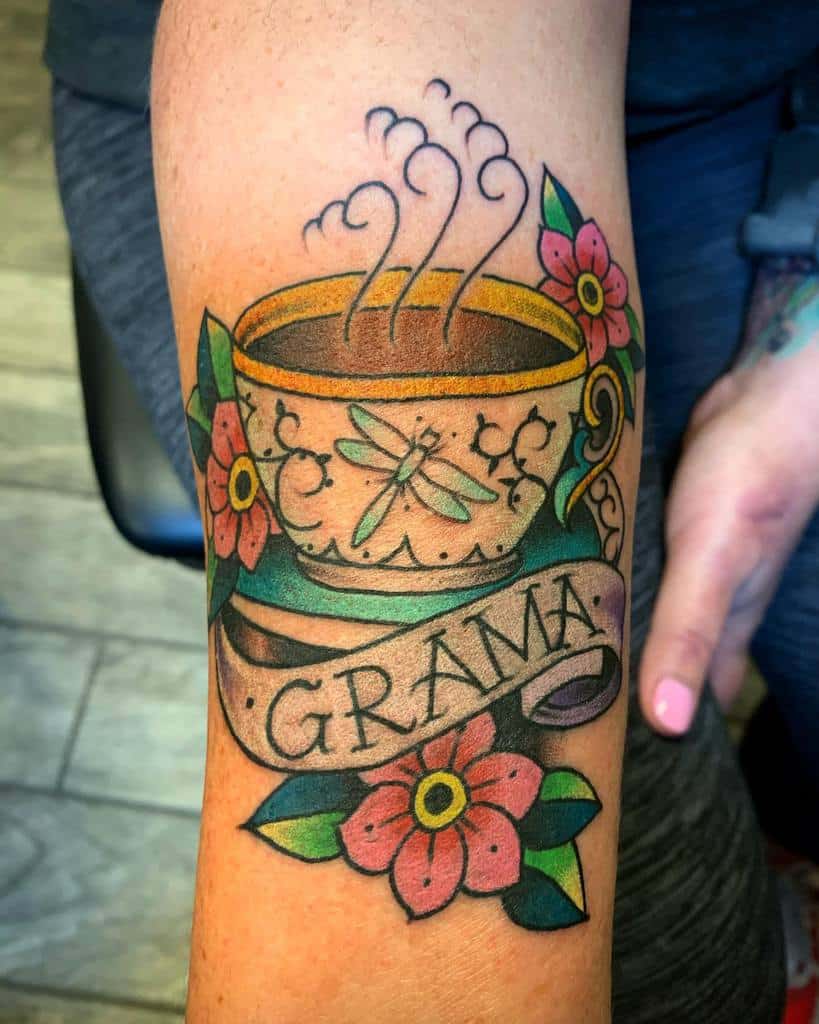 Colorful tattoo of a steaming teacup with floral patterns and a dragonfly design, surrounded by pink flowers, leaves and a banner reading "GRAMA"