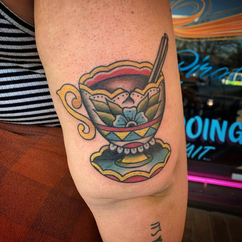 Colorful tattoo of an ornate teacup with a floral design, filled with a dark liquid, resting on a matching saucer with a spoon inside