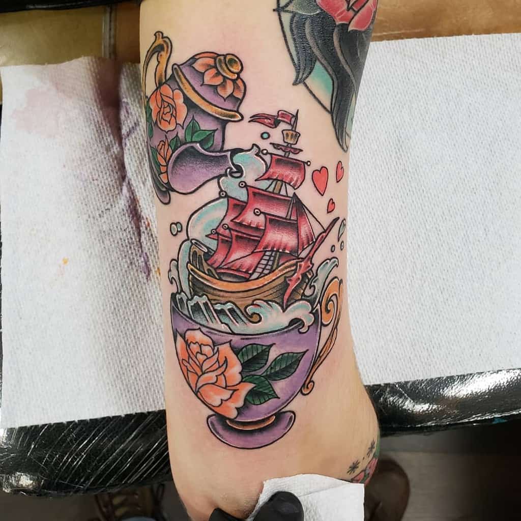Colorful tattoo of a floral teacup with ocean waves, a red sailing ship inside, and liquid pouring from a matching teapot, surrounded by hearts