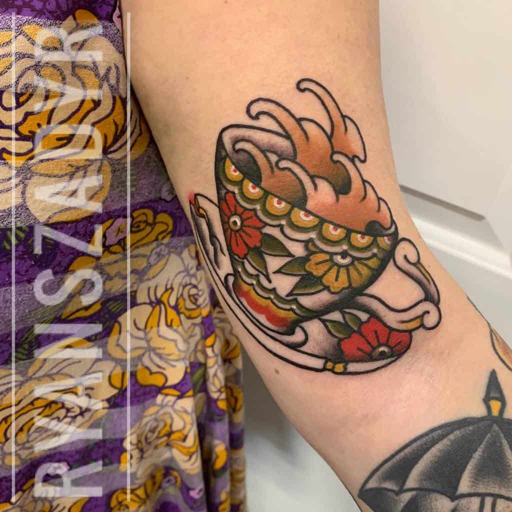 Colorful tattoo of a decorative teacup with floral patterns, spilling steaming liquid, with a matching saucer beneath