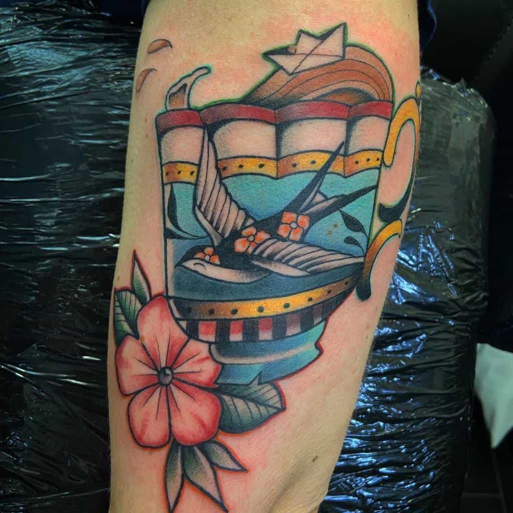 Colorful tattoo of a decorative teacup filled with a sea scene, featuring a swallow with three orange flowers on it, a paper boat on waves, and a large pink flower and leaves below