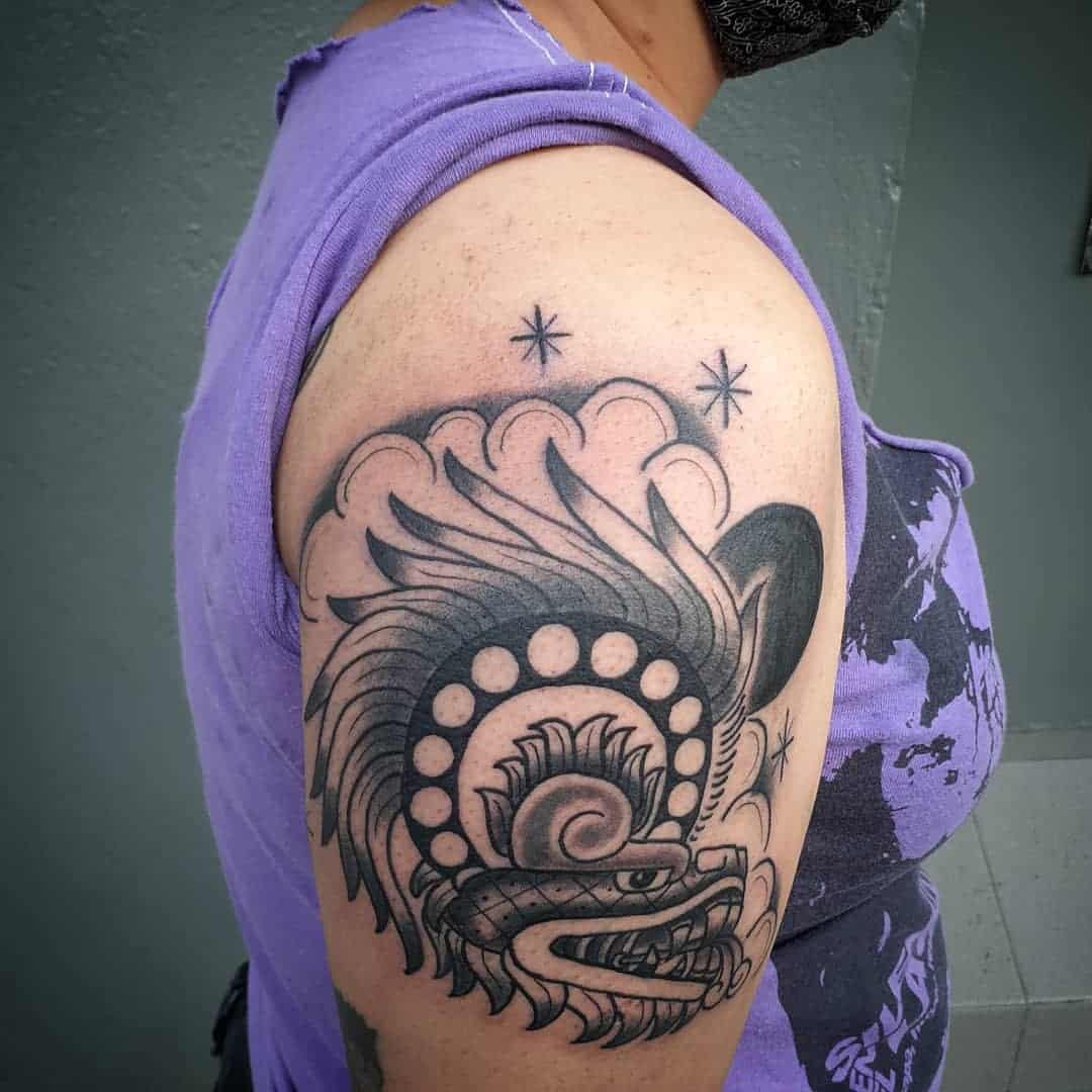 Upper arm tattoo of Quetzalcoatl in black and grey with intricate feathers and cloud details