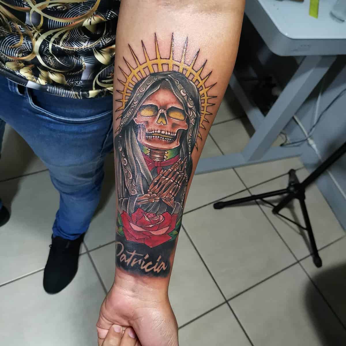 Vibrant Santa Muerte forearm tattoo featuring a radiant halo, red rose, and personalized name “Patricia”