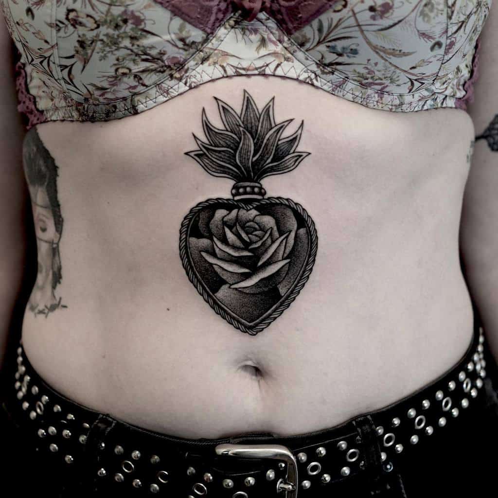 Black and gray Sacred Heart tattoo on the stomach, featuring a detailed rose inside and flame-like elements on top