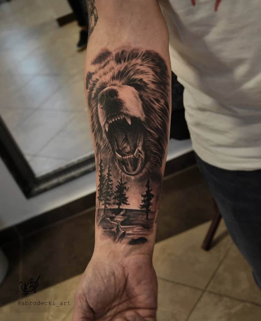 Realistic black and grey forearm tattoo of a roaring bear with a wilderness landscape below, featuring pine trees and a flowing river, symbolizing strength and nature.