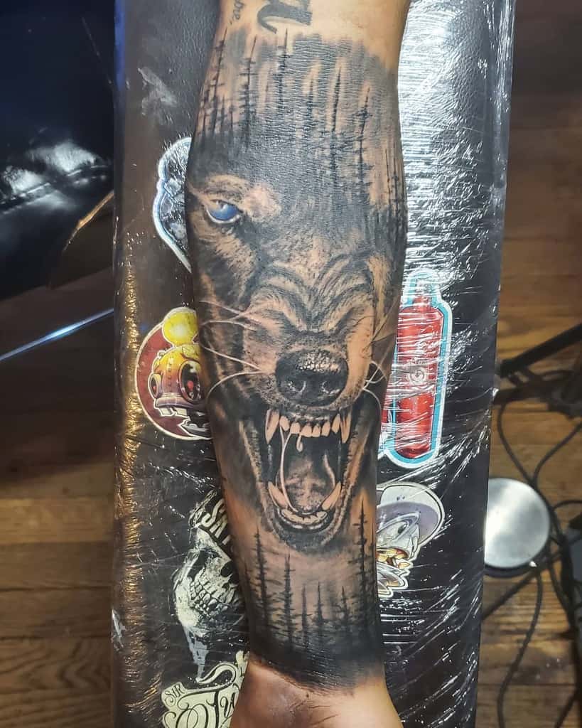 Forearm tattoo of a fierce, snarling wolf with blue eyes against a background of dark trees