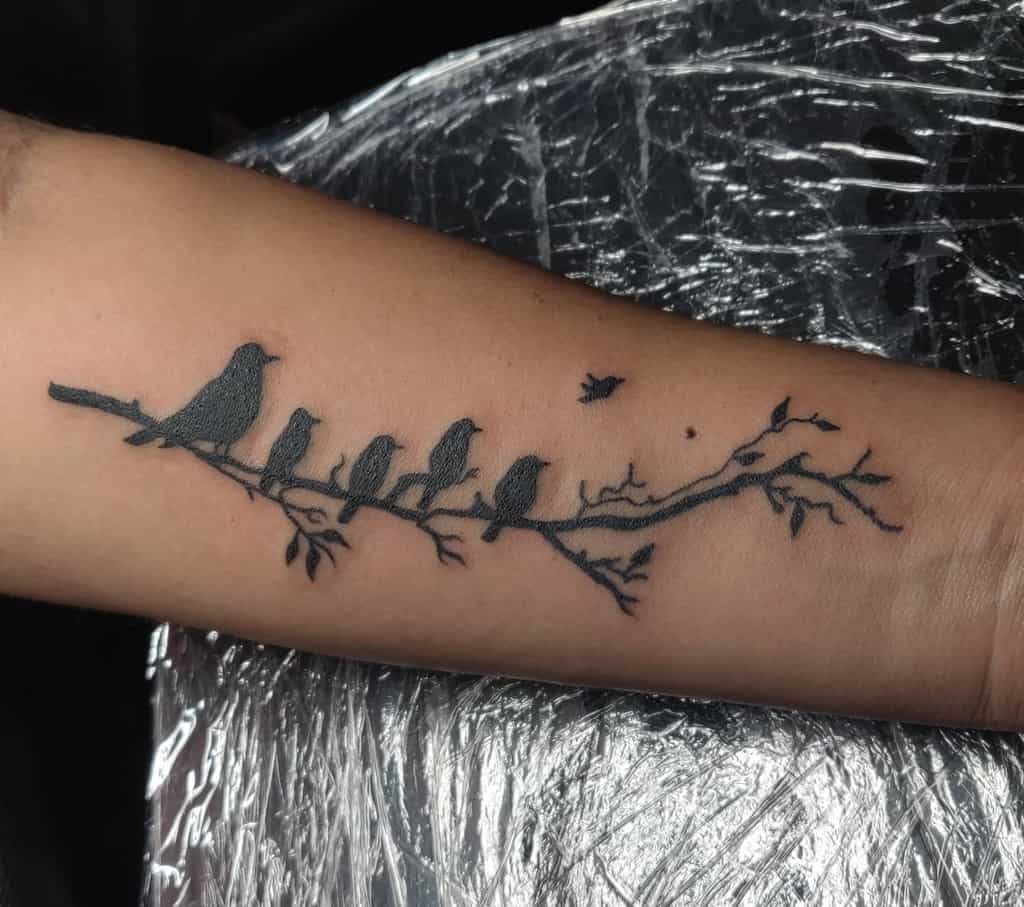 Tattoo of five birds perched on a delicate branch on an arm, with one bird flying away