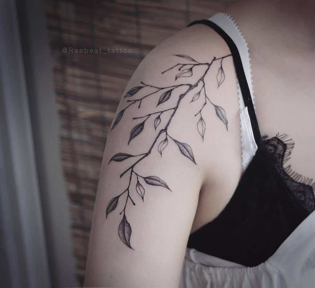 A person's shoulder adorned with a striking black and gray tattoo of an intricate branch and delicate leaves