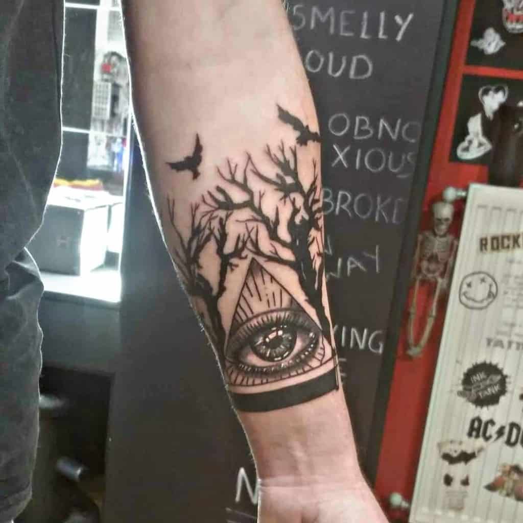 Forearm tattoo of an eye inside a pyramid, encircled by intricate tree branches and flying bats