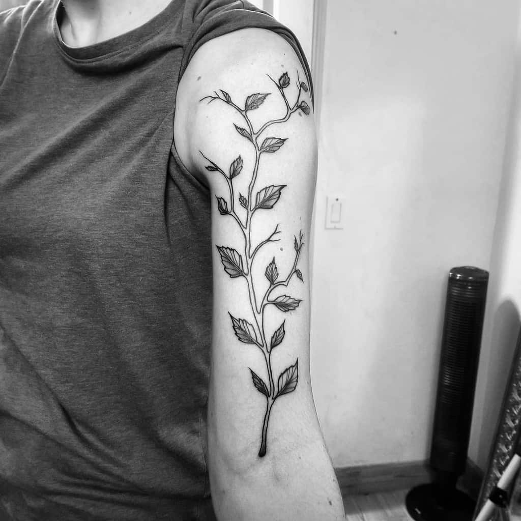 Black and white image of a person's arm adorned with a detailed botanical tattoo featuring leaves, buds, and an intricate branch
