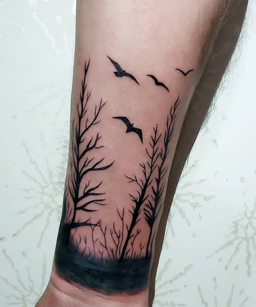 Arm tattoo of dark, leafless trees with intricate branches and flying birds