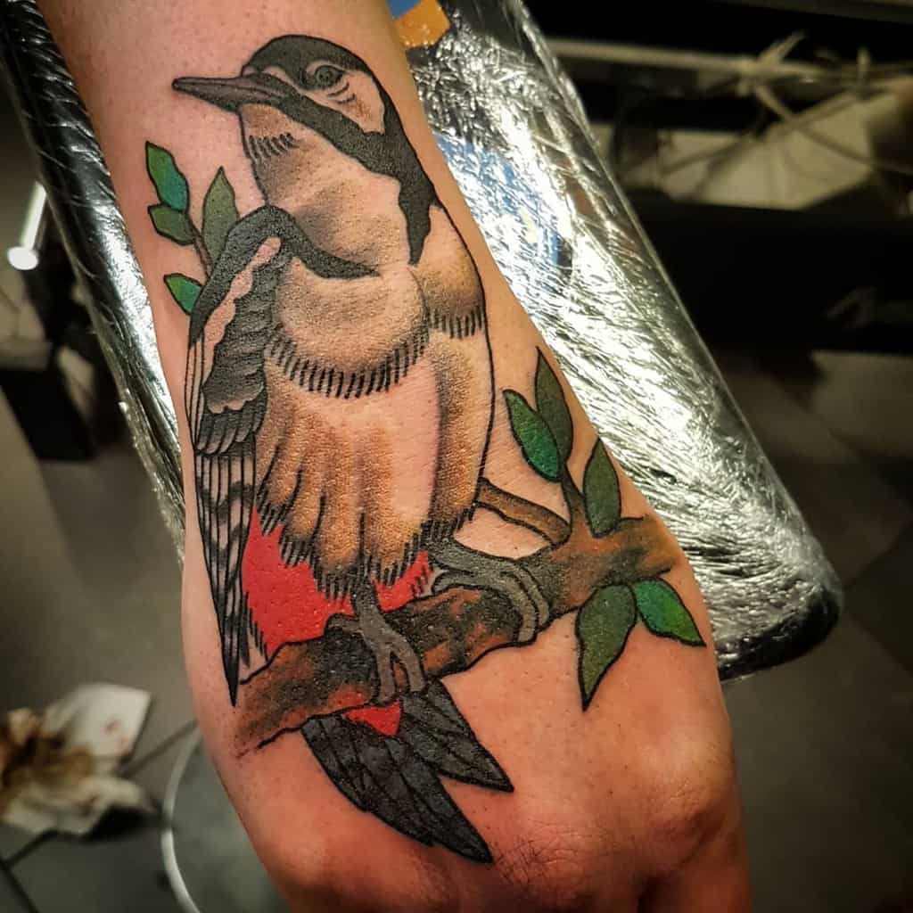 Colorful bird tattoo on hand, featuring a bird perched on a beautifully detailed branch with vibrant green leaves