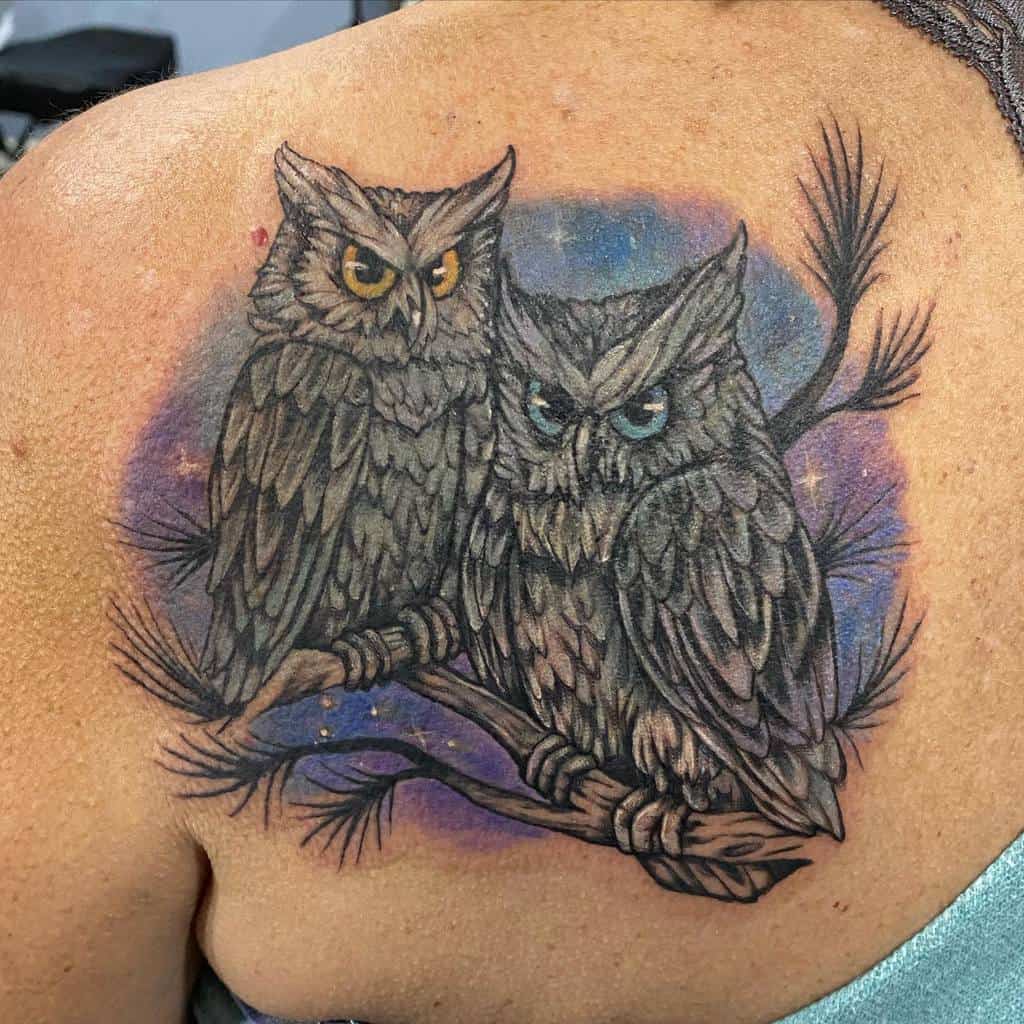Tattoo of two detailed owls gracefully perched on a branch, one with striking yellow eyes and the other with captivating blue eyes
