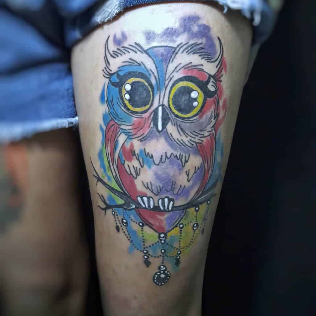 Colorful watercolor-style owl tattoo on a person's thigh, perched gracefully on a branch, boasting vibrant shades and detailed linework