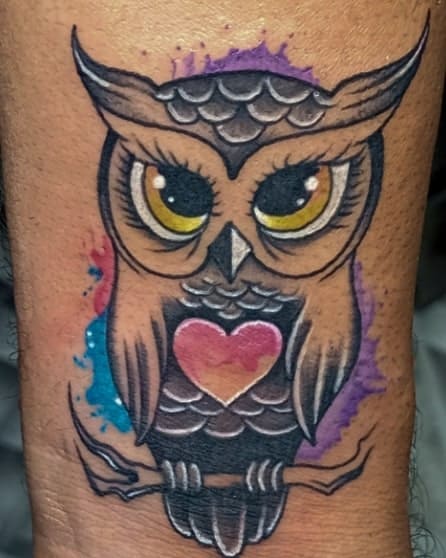 Colorful owl tattoo with large eyes and a heart on its chest, perched elegantly atop a detailed branch