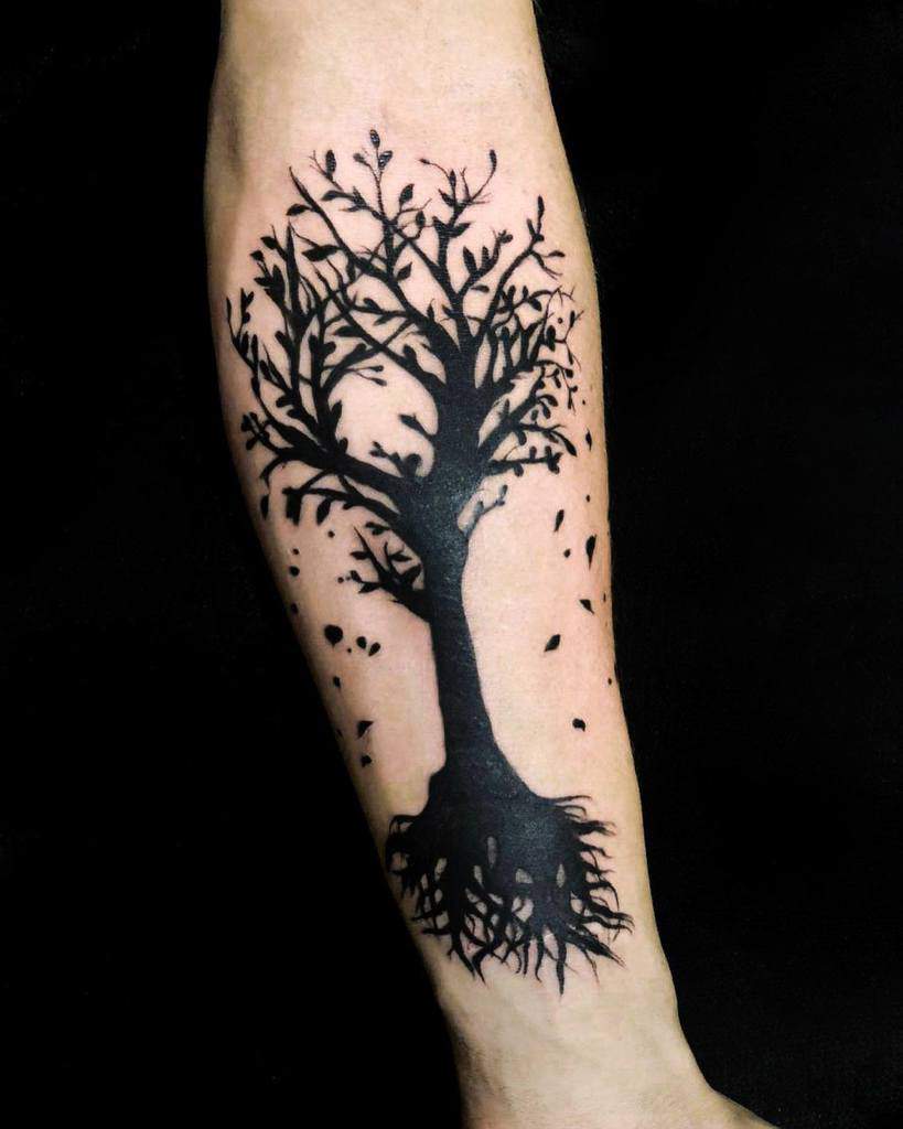 Forearm tattoo of a large black tree with branches, roots, and scattered falling leaves creating a striking silhouette
