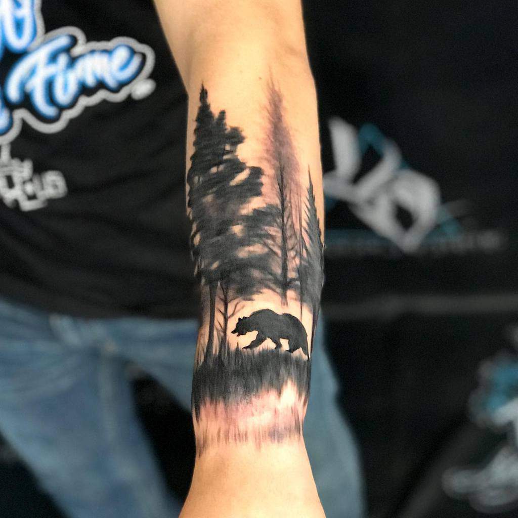 A forearm tattoo of a bear silhouette walking through a forest with tall trees