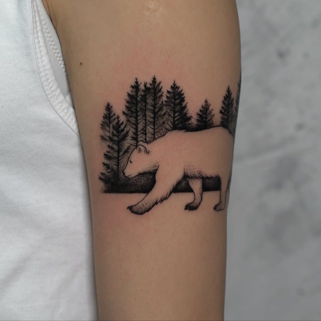 Tattoo of a bear with a forest silhouette, complete with towering trees, on an upper arm