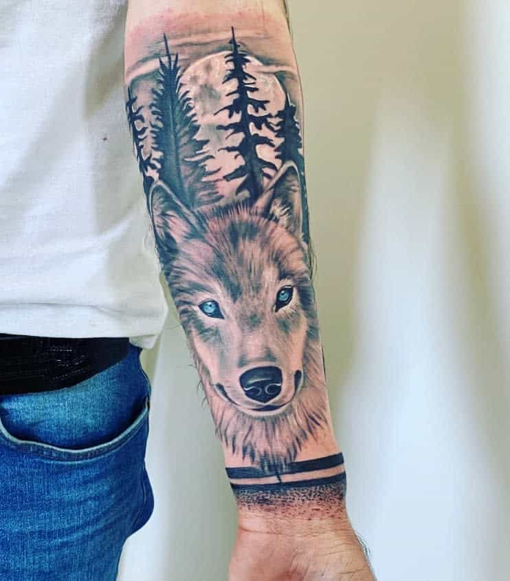 A forearm tattoo of a wolf with pine trees and a moon in the background