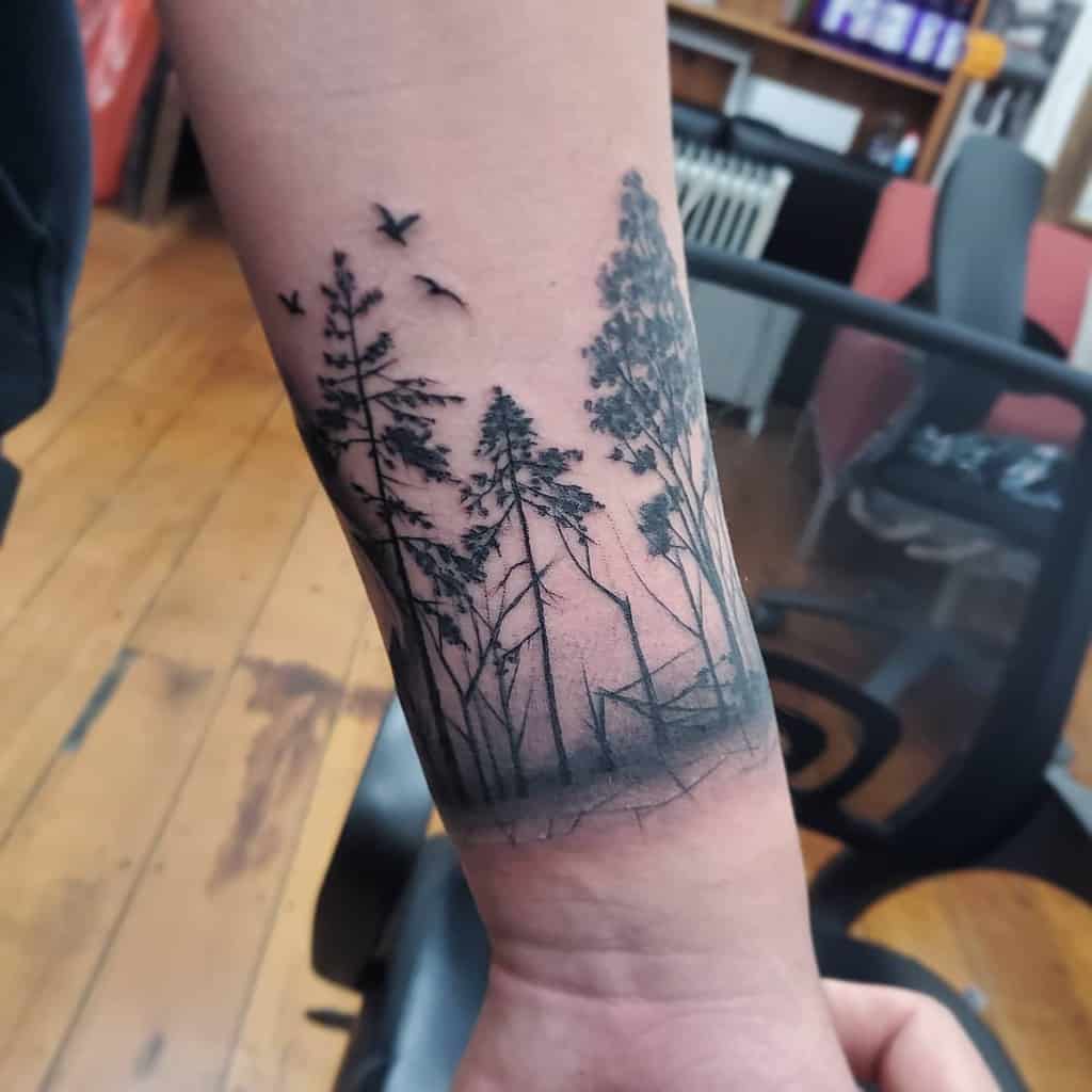 Wrist tattoo of tall trees with birds flying above, inked in black and grey
