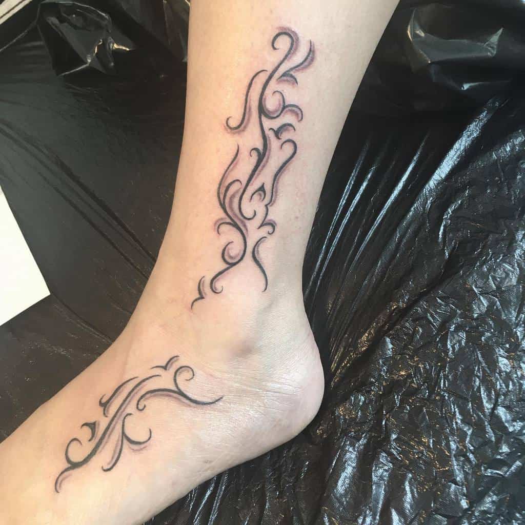 Black and gray ornamental tattoos on the side and ankle of a person's foot, with curved and swirling lines