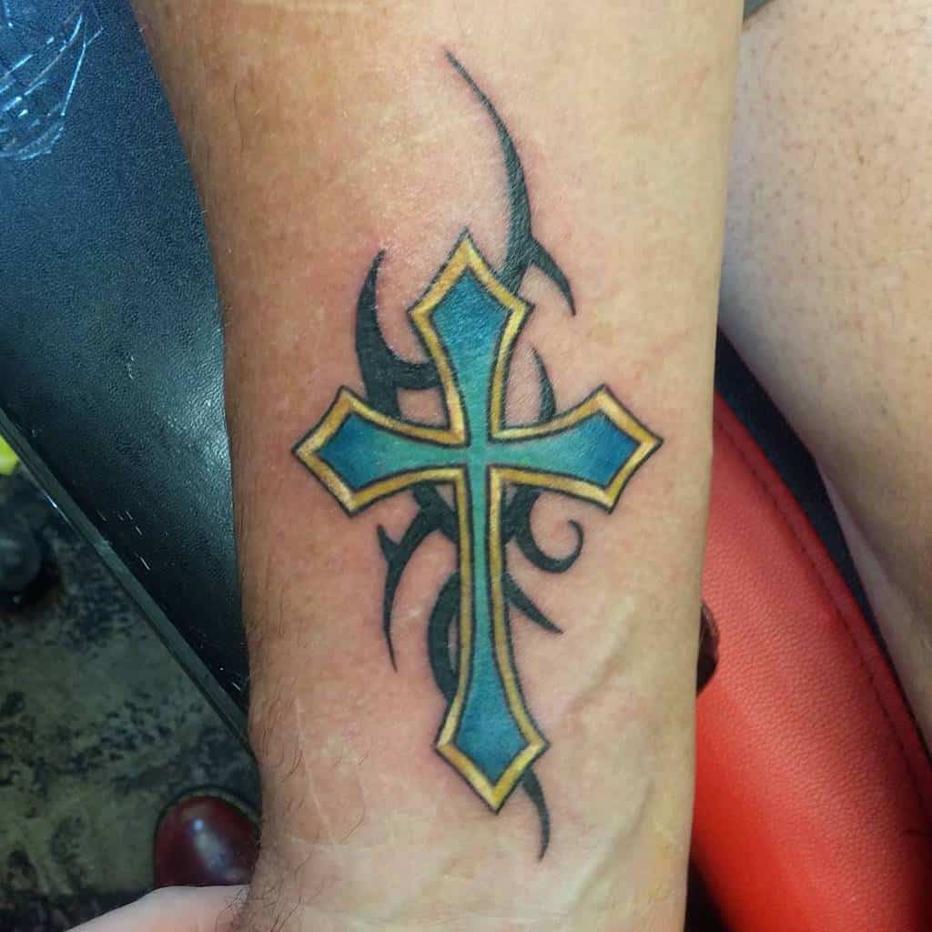 Tattoo of a blue cross with gold outline and intricate black tribal patterns elegantly adorning the forearm