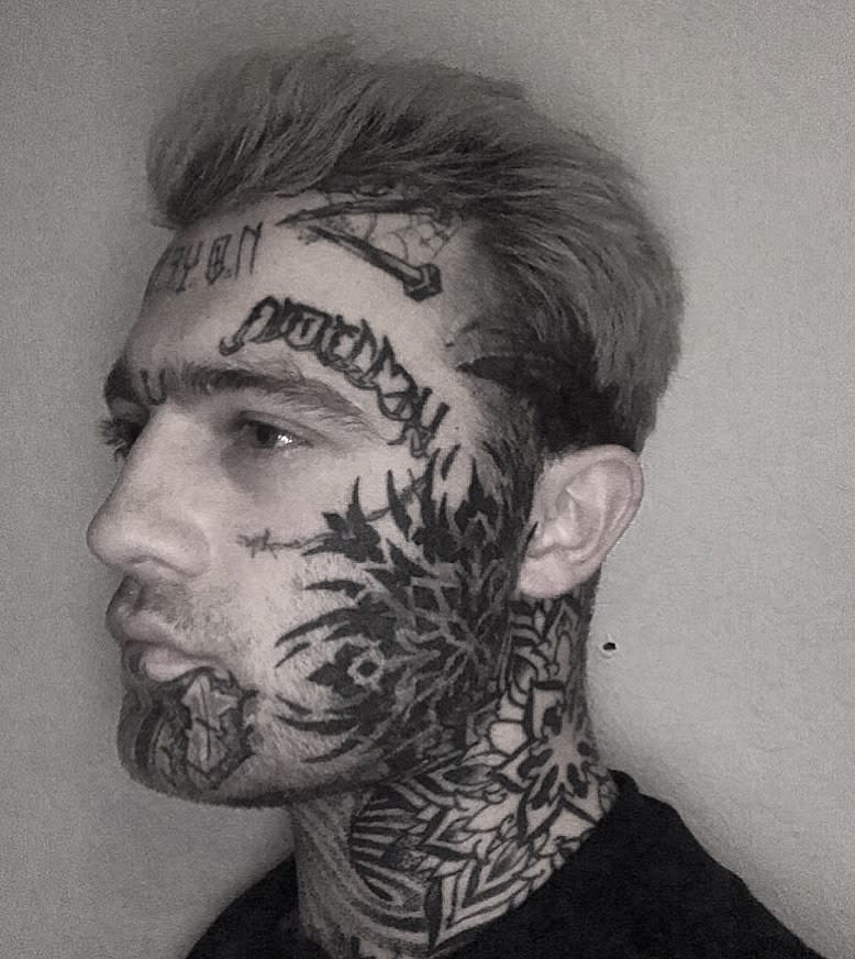 Side profile of a person with an extensive black tribal and script tattoo covering the face and neck, featuring bold, sharp patterns and text across the forehead and cheek