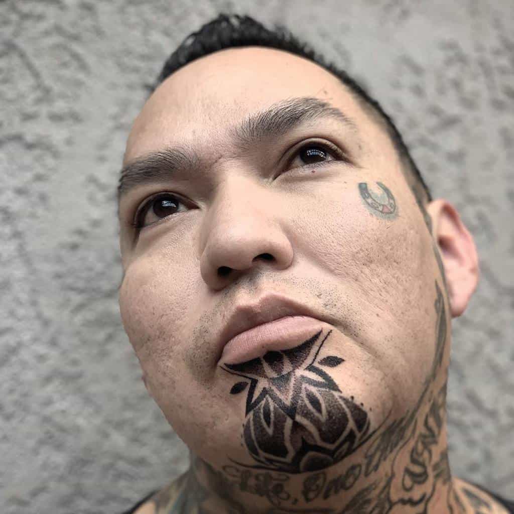 Bold, black tribal tattoo on the chin, featuring geometric shapes and sharp lines extending towards the neck