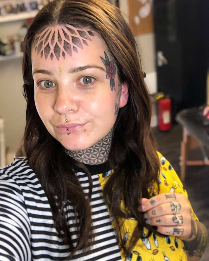Bold tribal tattoo on the forehead, featuring petal-like geometric shapes, along with floral and geometric designs on the side of the face, neck, and hands