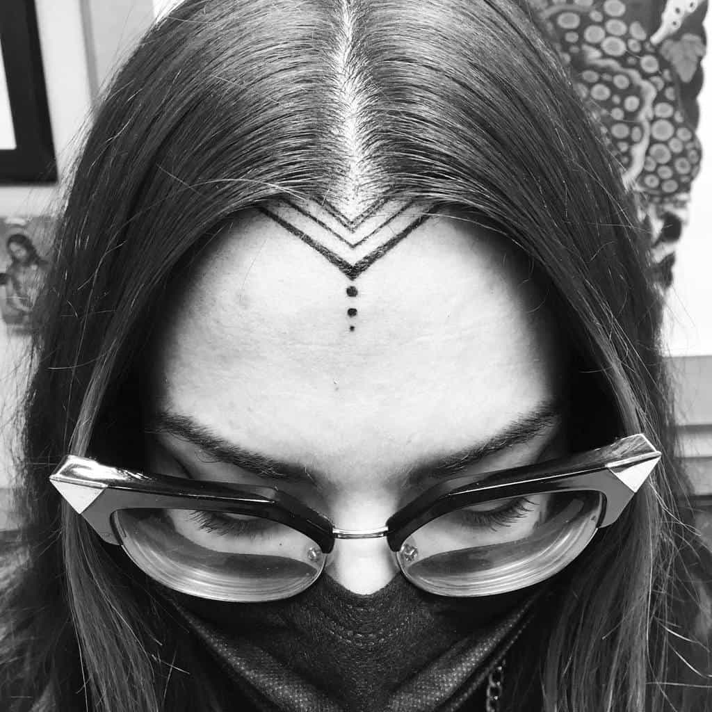 Minimalist tribal tattoo on the forehead, featuring sharp geometric lines forming a chevron shape, along with a vertical row of small dots