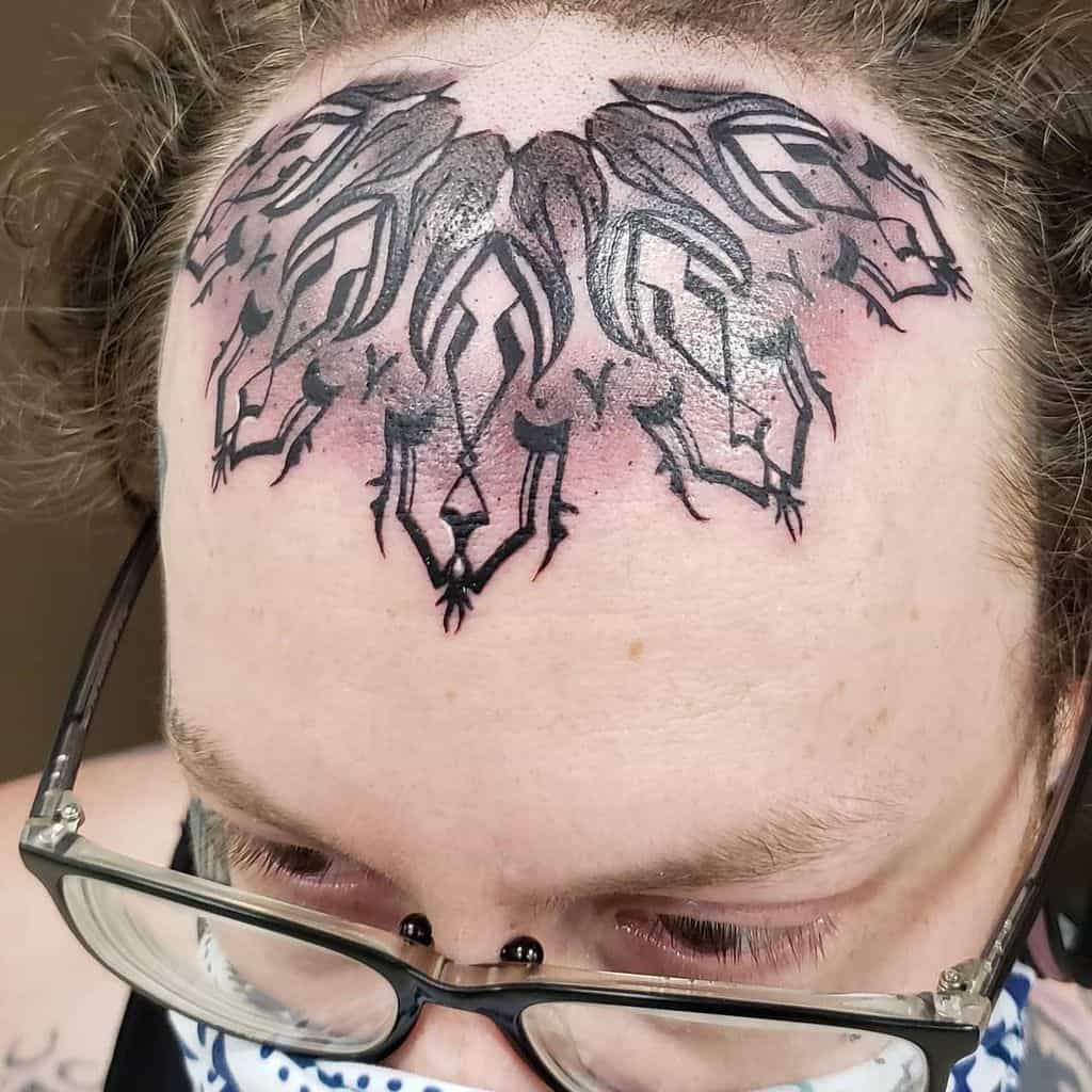 Bold, abstract tribal tattoo on the forehead, featuring intricate black lines and shapes, creating a symmetrical design with shaded elements