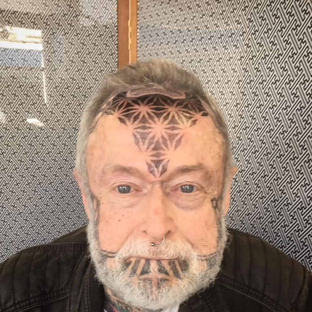 Geometric tribal tattoo covering the forehead, extending down the nose and chin, featuring symmetrical flower-like patterns and bold black lines