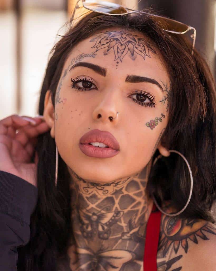 Tribal tattoo on the forehead, complemented by floral tattoos on the cheek, small dot accents, and a large intricate tattoo covering the neck and chest.