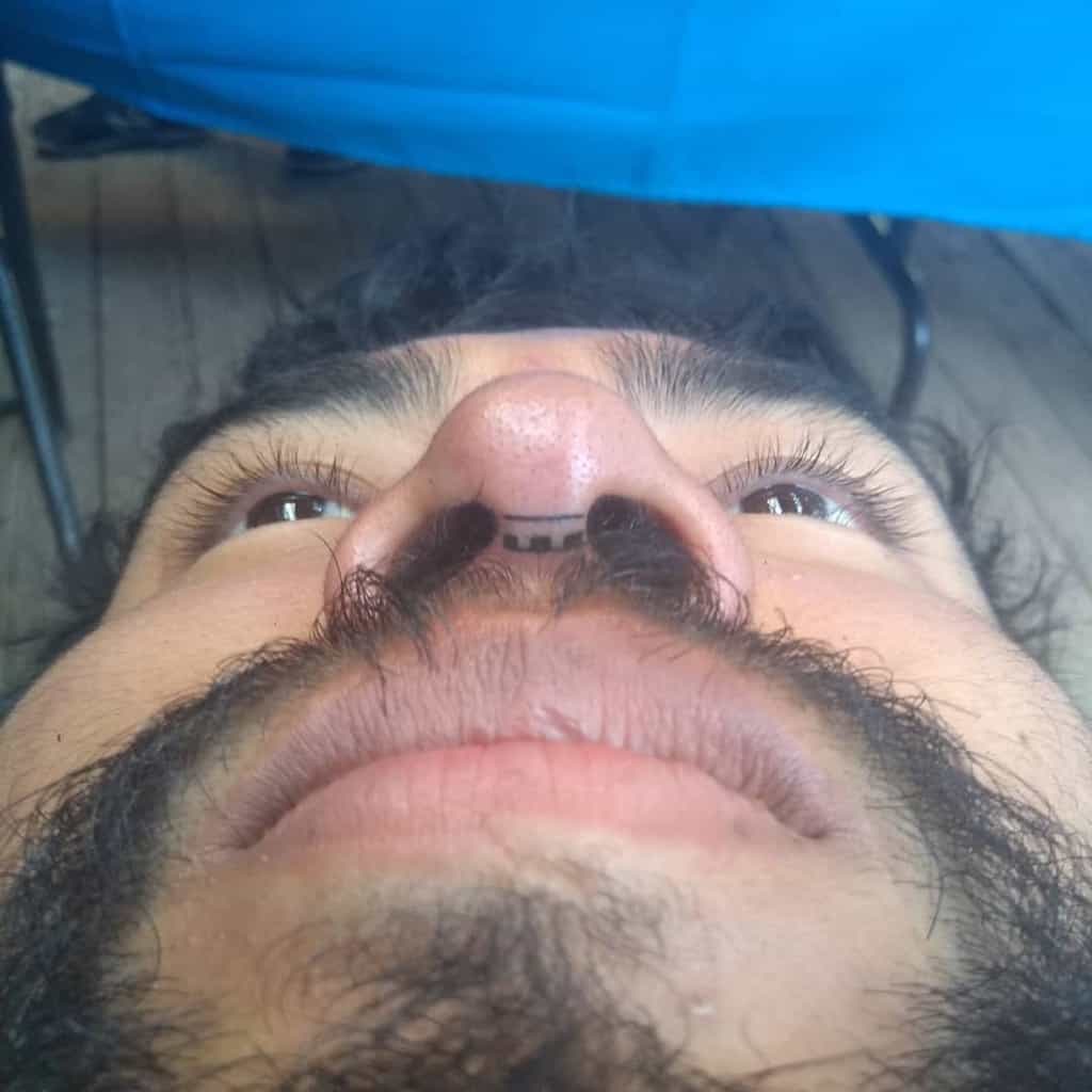 Small, minimalist tribal tattoo on the underside of the nose, featuring simple horizontal and vertical lines