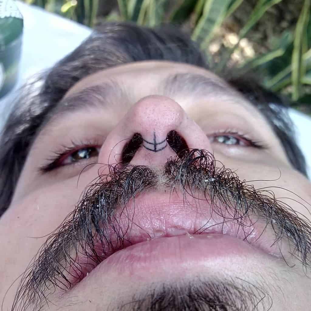 Man's nose tattoo featuring a small symmetrical design between the nostrils, with a visible mustache