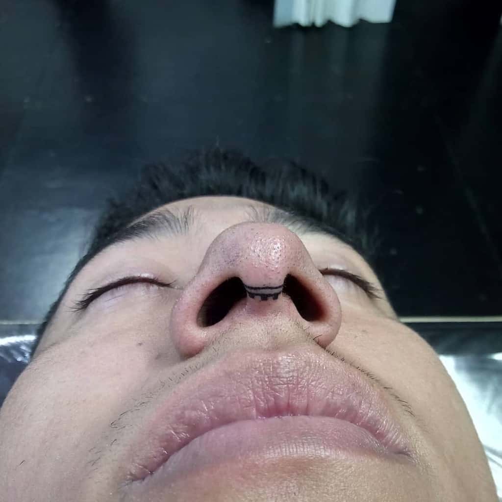 Man's nose tattoo with a small, intricate symmetrical design beneath the nostrils, featuring clean, thin lines